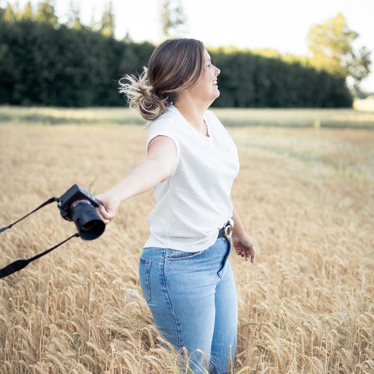 Anne Merz Fotografin mit Kamera in einer Weizenfeld, die glücklich die Arme ausbreitet.